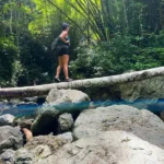 Teresa Prendes-Walls hiking across a log over rocks at Charco Prieto waterfall in Puerto Rico during Root to Rise Retreat